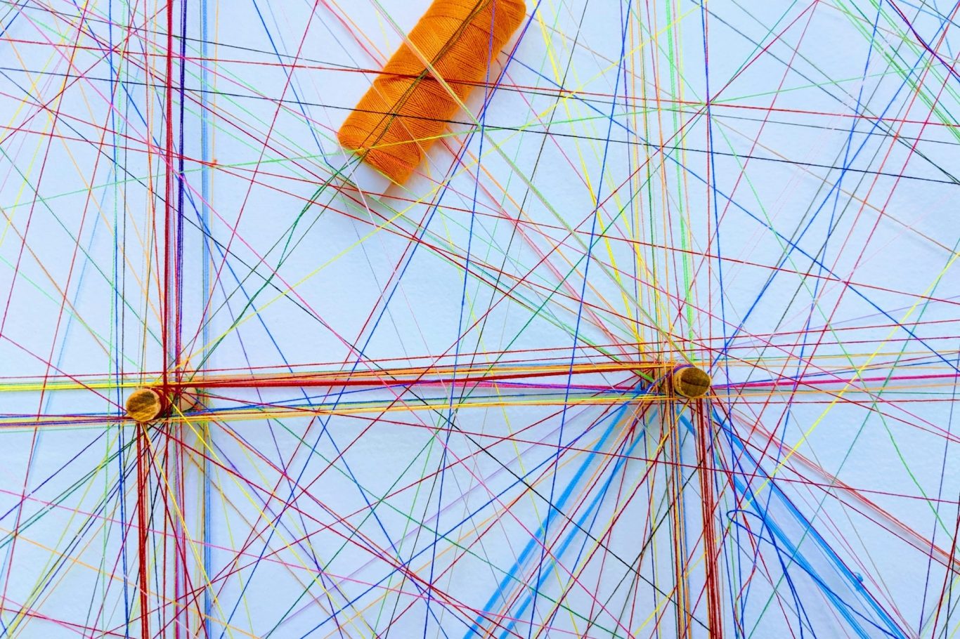 Kooperation Bunte Fäden kreuzen sich auf einer weißen Fläche, mit einer orangefarbenen Garnrolle.