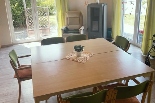 Wohngemeinschaften Holztisch mit Stühlen, Pflanze in der Mitte, helles Zimmer mit großen Fenstern und Gartenblick.